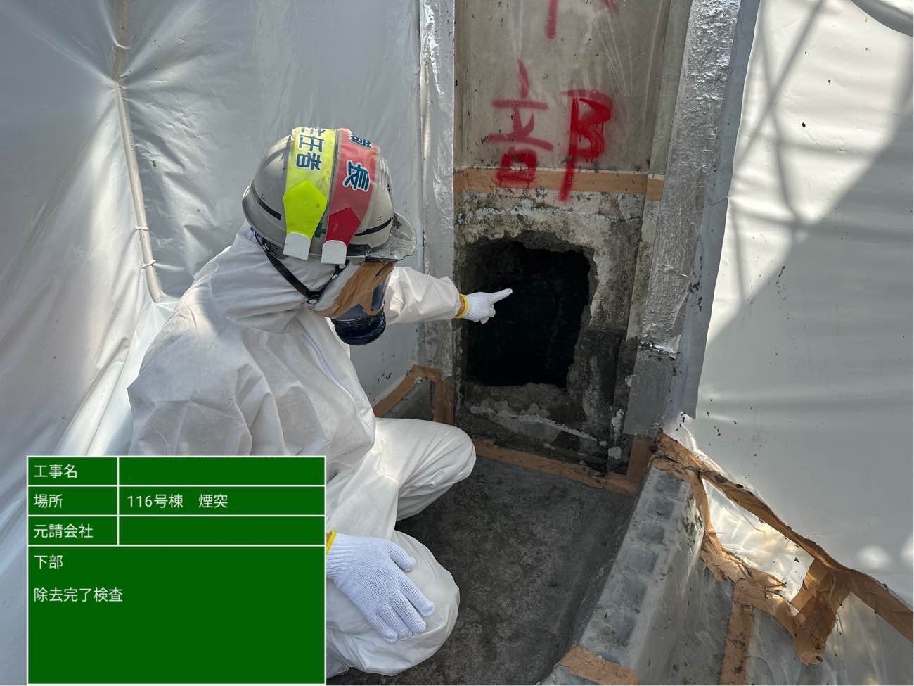 防護服を着た作業責任者が煙突下部を指さし、除去完了後の状態をチェックしている様子